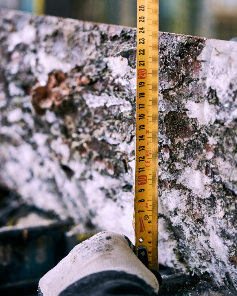 Gloved hand measuring a frost-covered spruce slab with a tape at the sawmill—sizing tonewood billets for acoustic guitars.