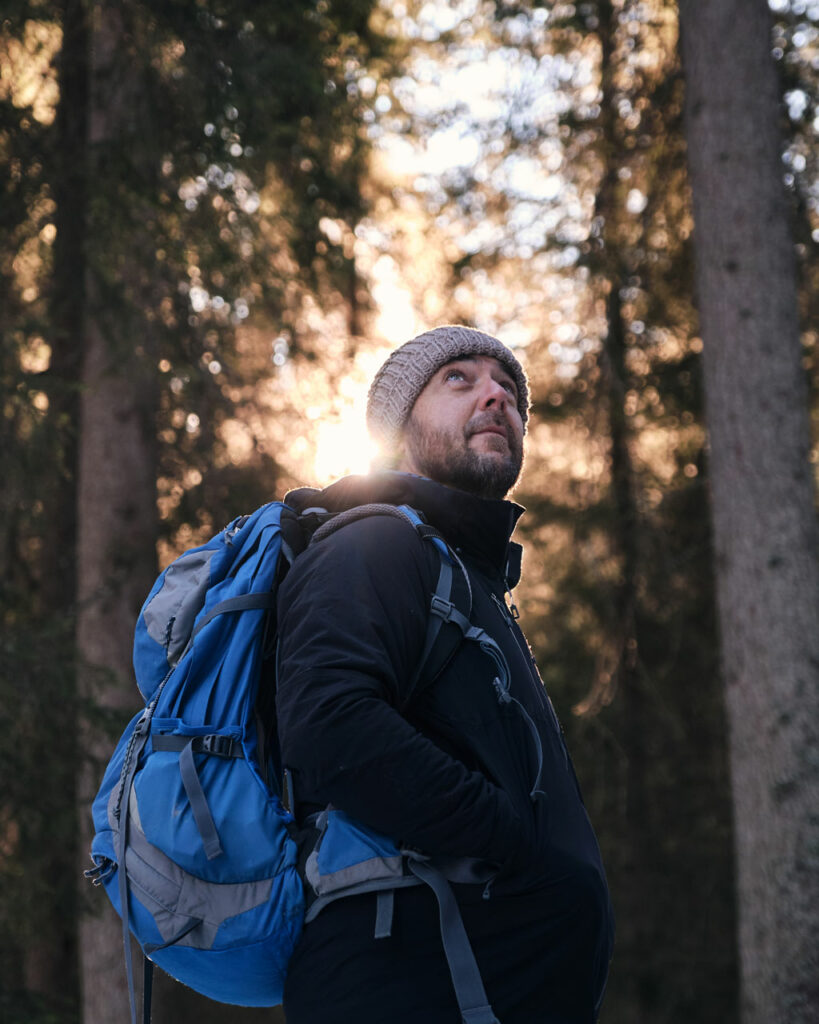 Thom Orgler, South Tyrolean luthier, looking up in a Dolomite forest at sunrise, wearing a blue backpack while scouting tonewood.