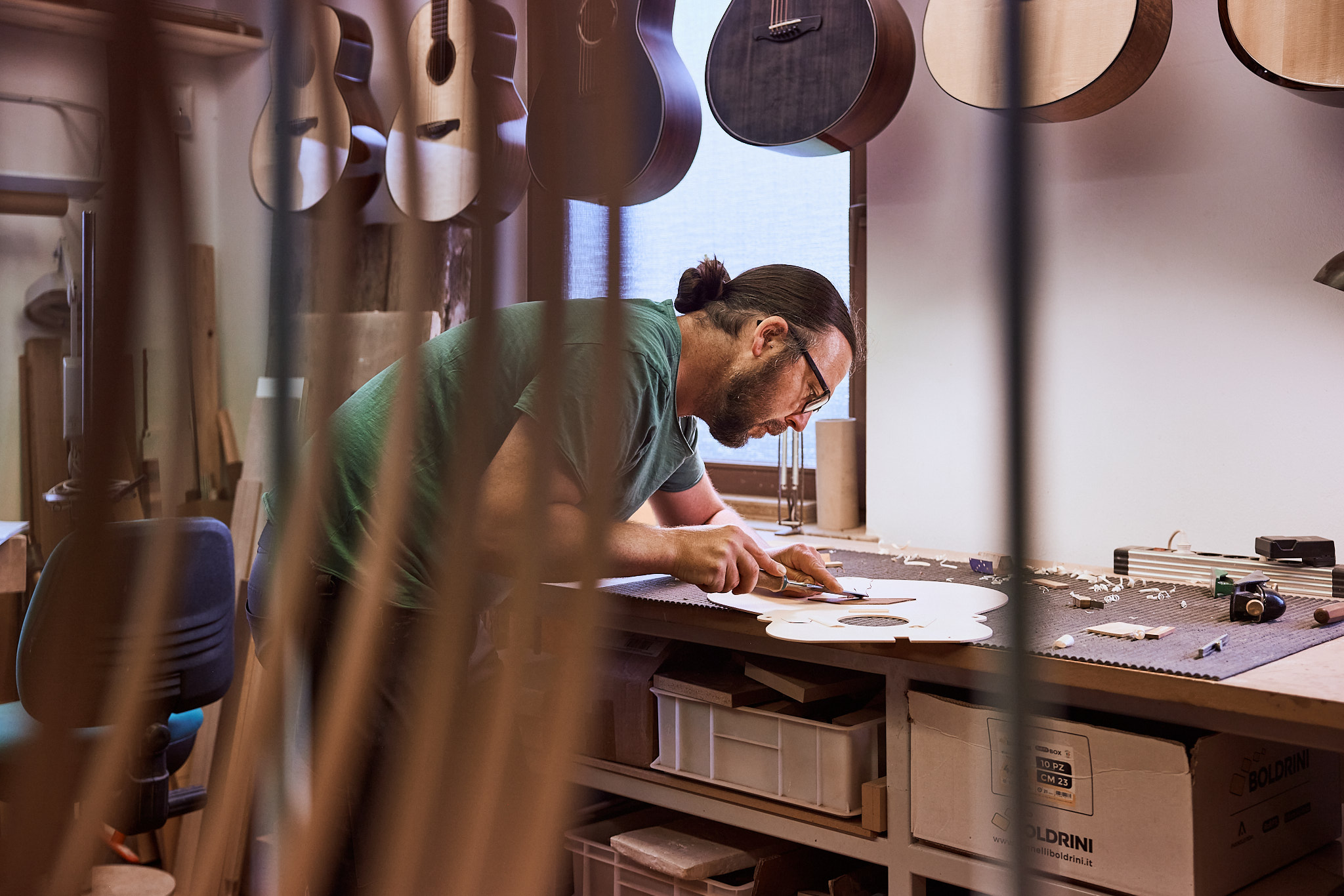 Thom Orgler, South Tyrolean luthier, carving an acoustic guitar soundboard at his workshop bench, with finished guitars hanging behind.