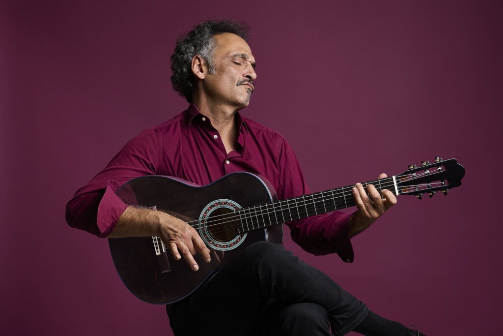 Chitarrista suona una chitarra classica Thom Orgler Enrosadira Cypress con finitura scura Smokegrain davanti a uno sfondo bordeaux.