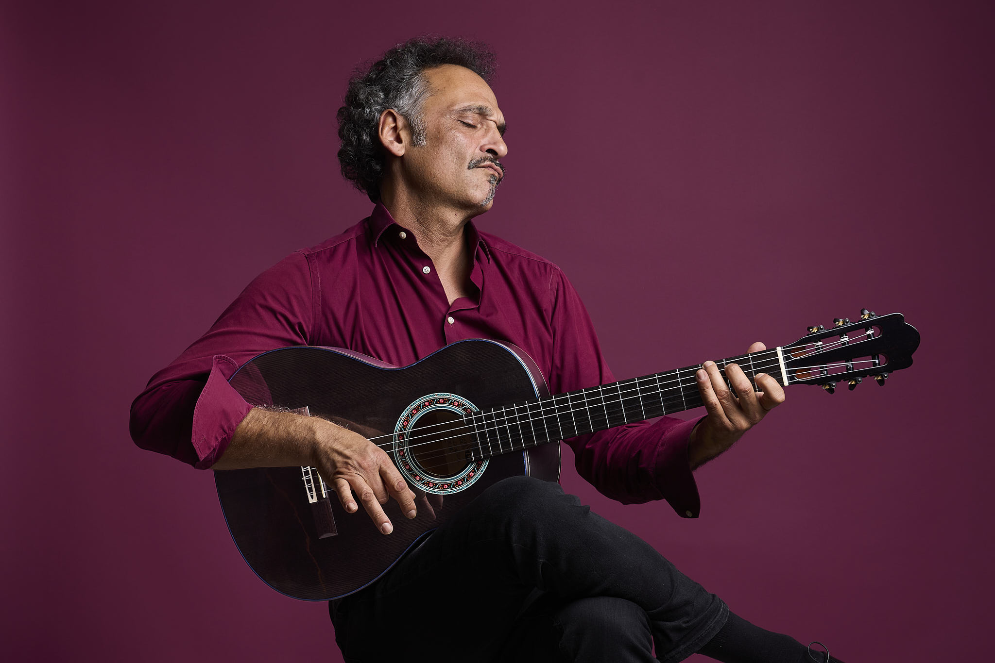 Guitarist playing a Thom Orgler Enrosadira Cypress classical guitar with a dark Smokegrain finish against a burgundy studio background.