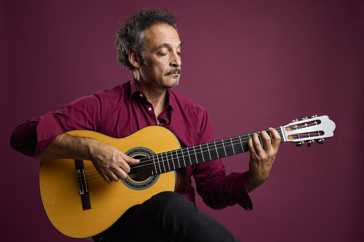 Guitarist playing a Thom Orgler Enrosadira classical guitar with nylon strings against a burgundy studio background.
