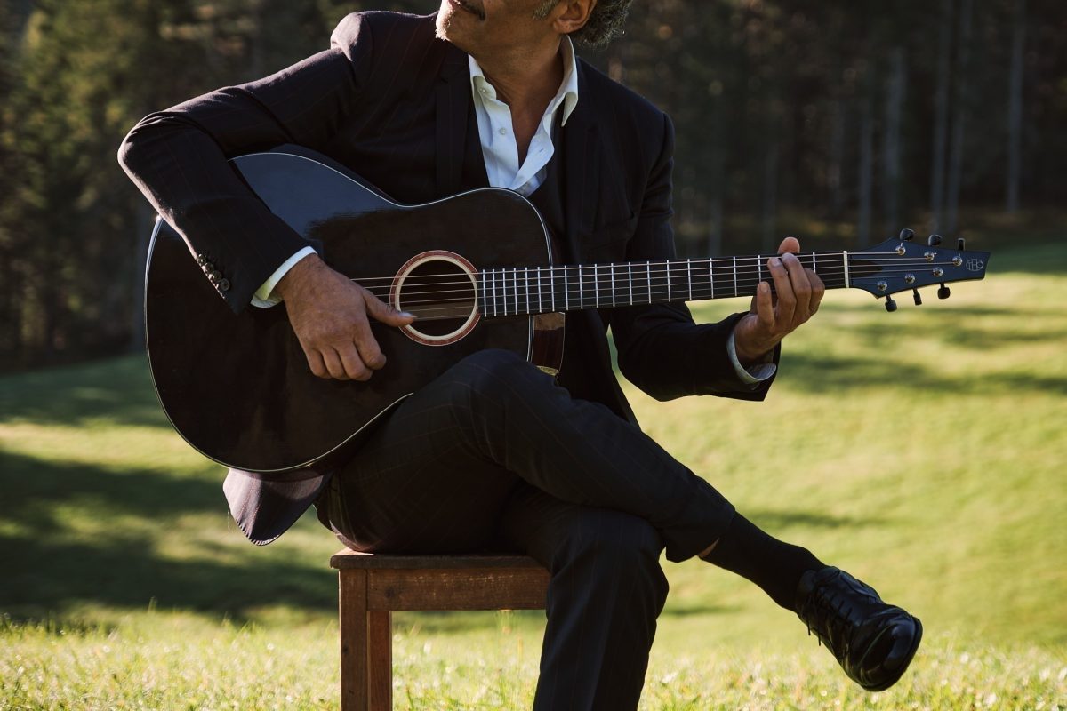 Manuel Randi performing on his Thom Orgler Latemar (PU high-gloss Smokegrain finish), sitting on a wooden chair in a green field.